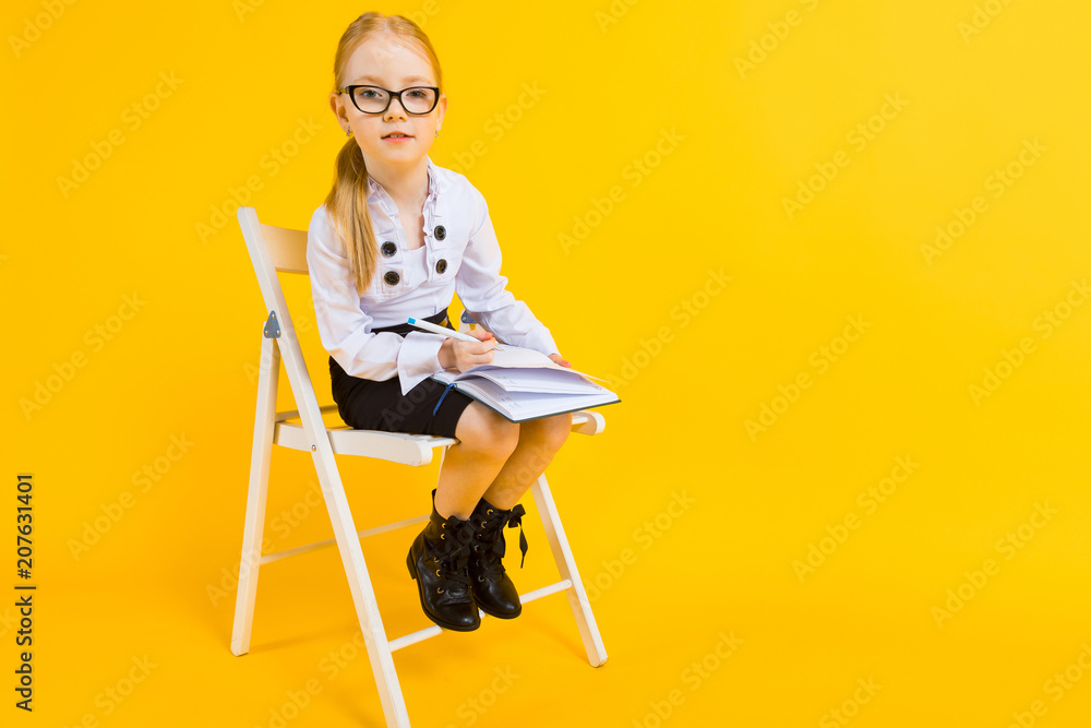Fototapeta premium Girl with red hair on a yellow background. A charming girl in transparent glasses sits on a white chair and makes notes in a notebook.