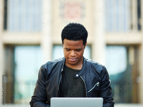 Portrait of concentrated African American student in leather jacket studying on his laptop outdoors © Comeback Images