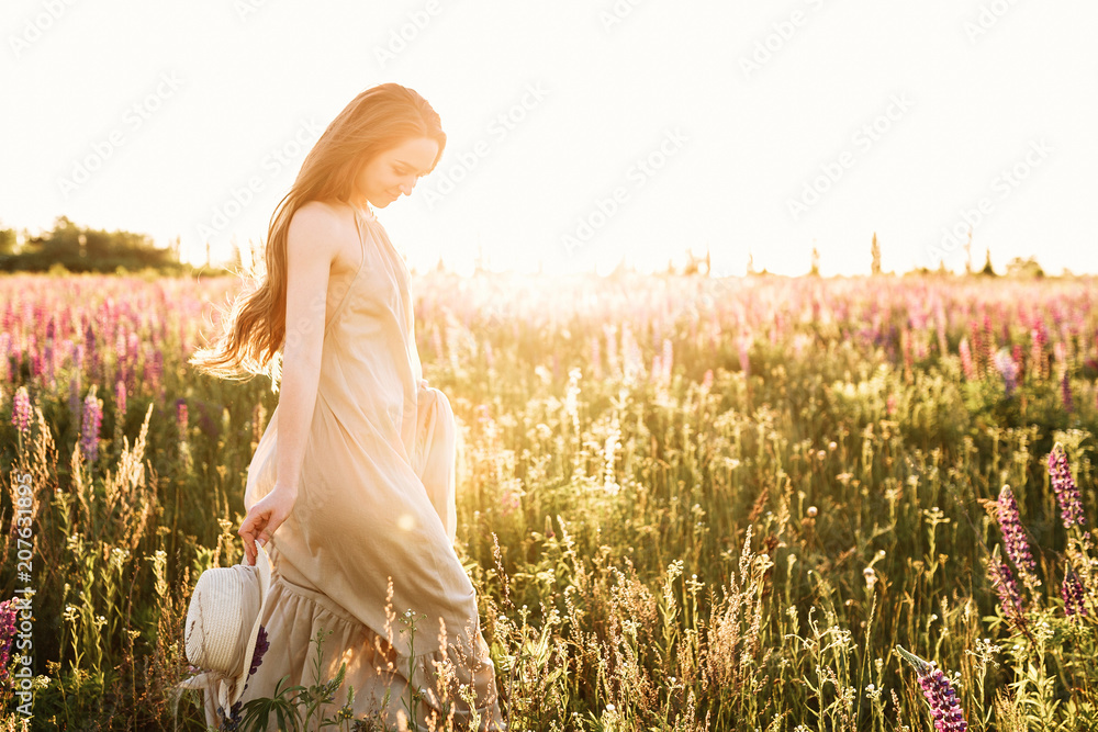 Obraz premium Young woman walking in lupine flower field with sunrise on the background. Warm orange sunbeam light