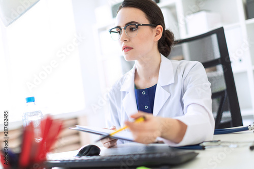 A beautiful young girl in a white robe is sitting at a computer desk with documents and a pen in her hands. Photo with depth of field, focus on girl. photo