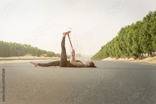 Athletic white female fitness model wearing long black sports wear with does a pilates exercises with rubber band in the middle of a quiet street with sun flare 
