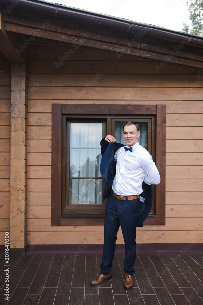 Morning of the groom. The groom smiles in a blue suit and a bow tie. Outdoor. A wooden brown house. Morning of the groom. The groom smiles in a blue suit and a bow tie. Outdoor. A wooden brown house.