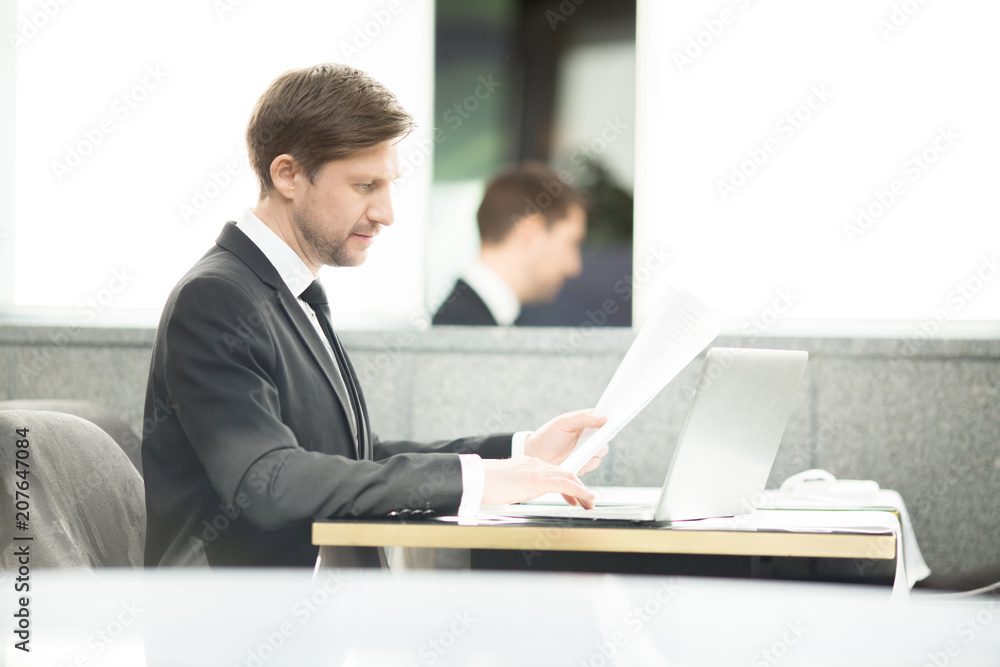 Young analyst or financier with papers working in front of laptop by table Young analyst or financier with papers working in front of laptop by table
