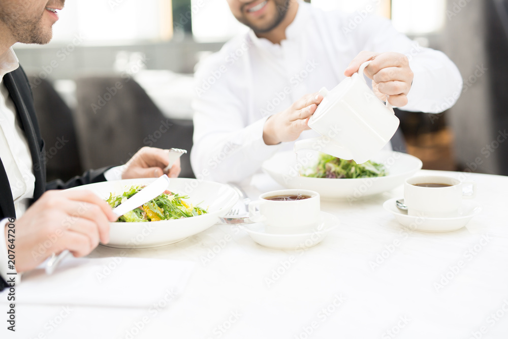 One of business partners pouring hot tea from teapot into cup during business lunch