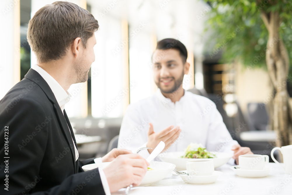 Businessman with knife and fork eating lunch in restaurant and listening to business partner