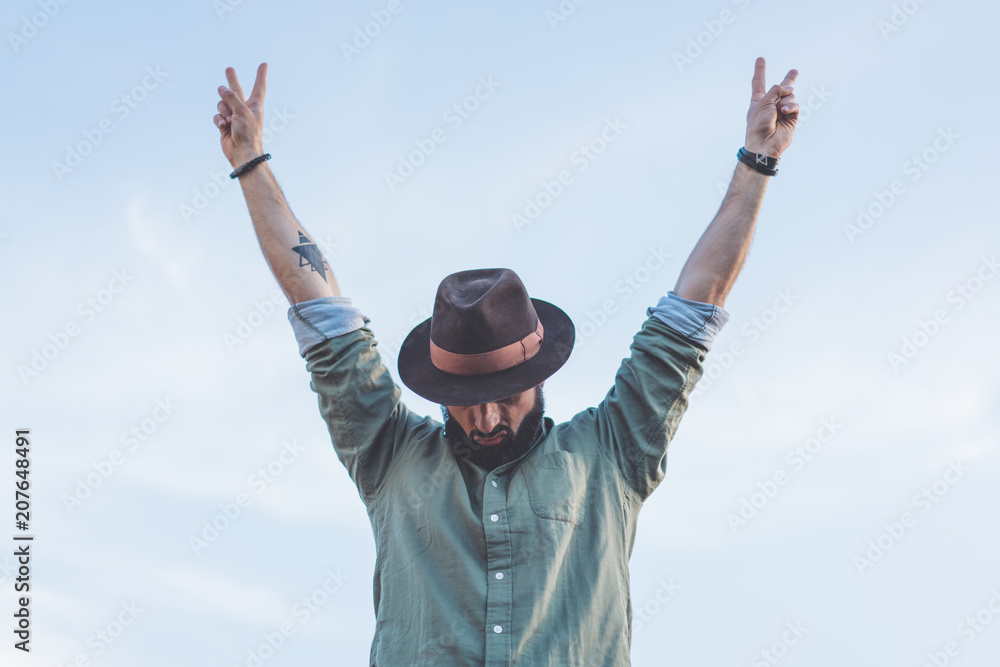 Bearded Hipster male wearing fashion hat, enjoying view of blue and clear sky with raised his hands up victory symbol Bearded Hipster male wearing fashion hat, enjoying view of blue and clear sky with raised his hands up victory symbol