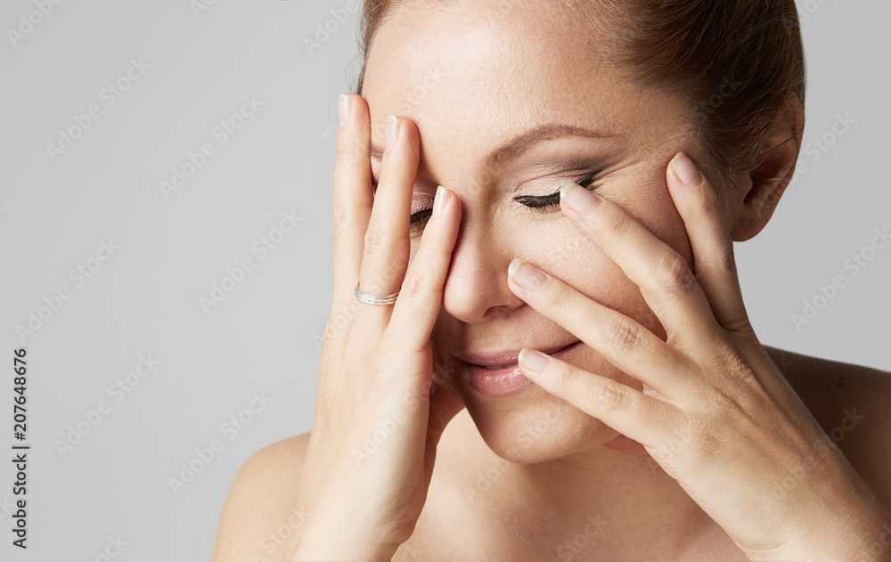 Beautiful smiling woman with clean skin, natural make-up, and closed eyes touching hands her face on gray background. Close up portrait Beautiful smiling woman with clean skin, natural make-up, and closed eyes touching hands her face on gray background. Close up portrait