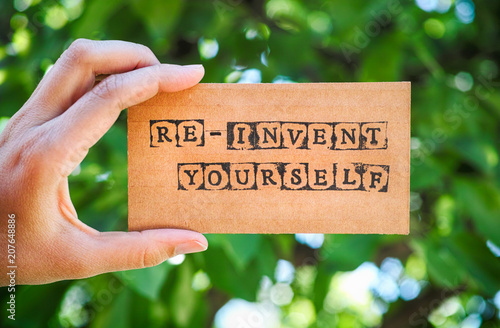 Woman hand holding cardboard card with words Re-Invent Yourself against green nature background.