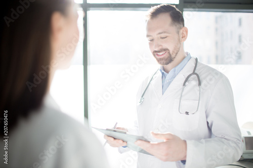 Happy young doctor reading medical notes while communicating with his colleague or assistant