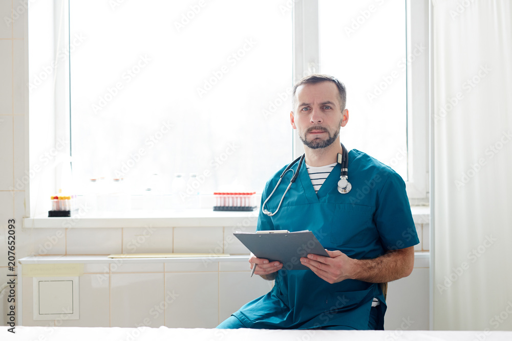 Serious doctor in uniform sitting by window with clipboard during work in clinics Serious doctor in uniform sitting by window with clipboard during work in clinics