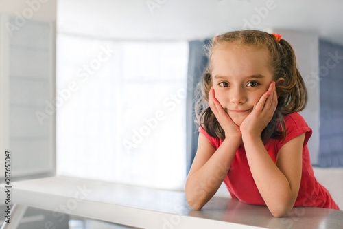 Pretty little girl in pink t-shirt and with pigtails leaning on hands at table and looking at camera