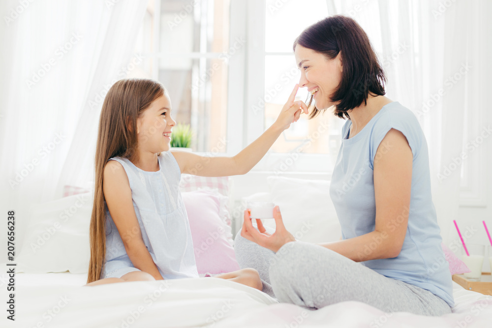Pretty small female child has fun together with her mother, applies beauty cream on her nose, sit together on comfortable bed, pose in spacious light bedroom. People, happiness and family concept