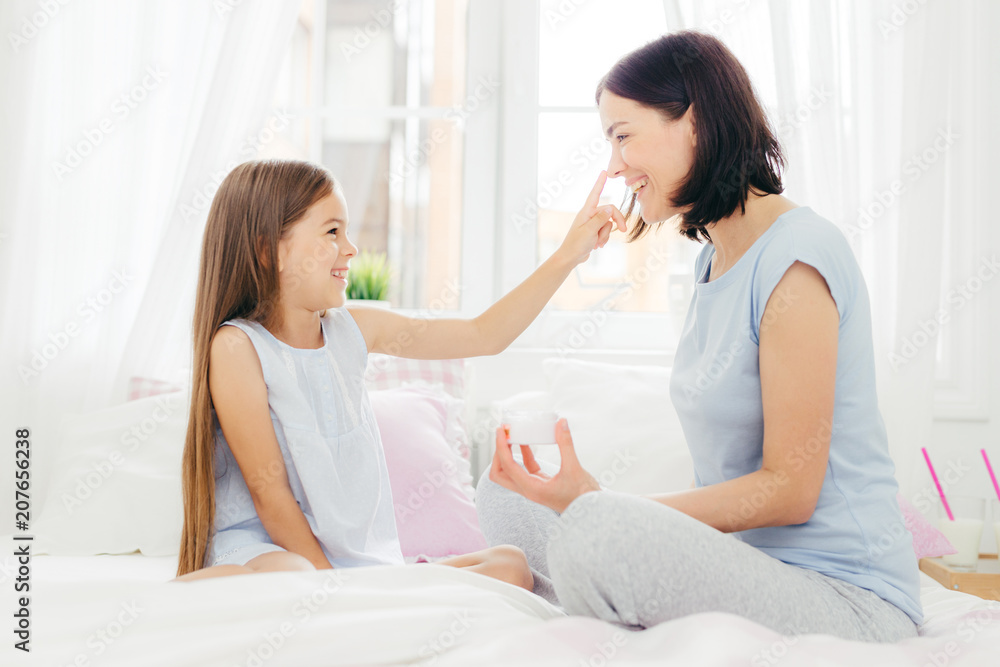 Pretty small female child has fun together with her mother, applies beauty cream on her nose, sit together on comfortable bed, pose in spacious light bedroom. People, happiness and family concept Pretty small female child has fun together with her mother, applies beauty cream on her nose, sit together on comfortable bed, pose in spacious light bedroom. People, happiness and family concept