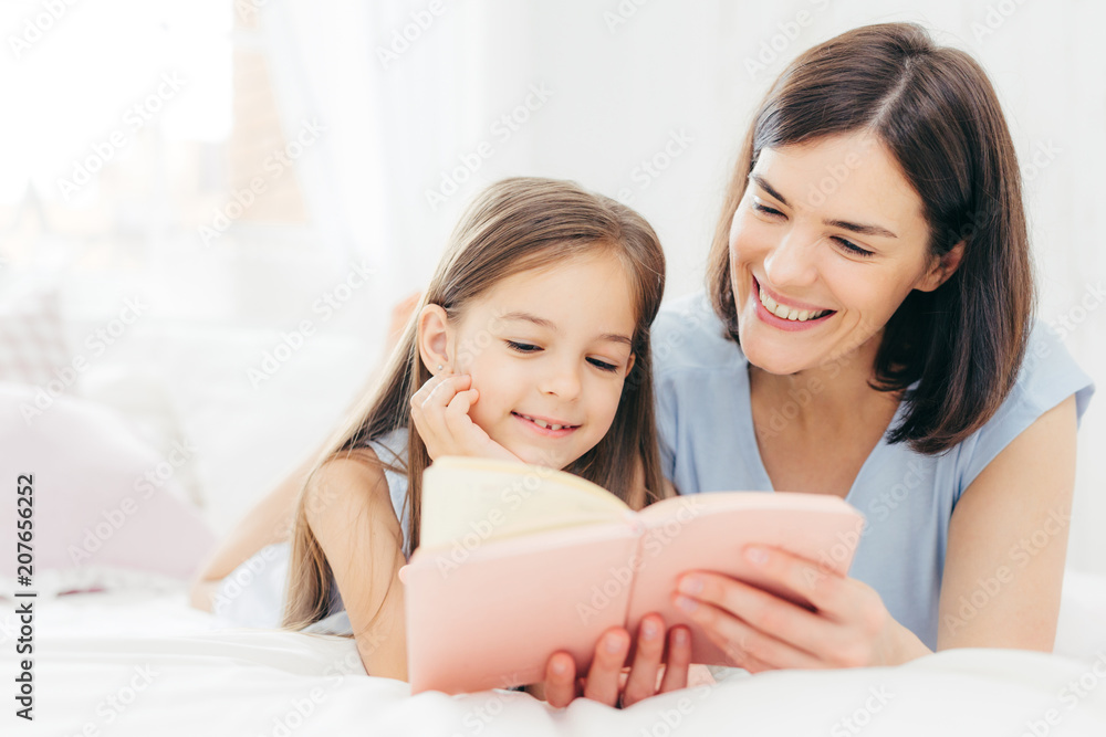 Pretty female mother and her small daughter read interesting book in bedroom, lie on comfortabled bed during morning. Lovely mum reads fairty tale for her little female kid, who is very curious Pretty female mother and her small daughter read interesting book in bedroom, lie on comfortabled bed during morning. Lovely mum reads fairty tale for her little female kid, who is very curious