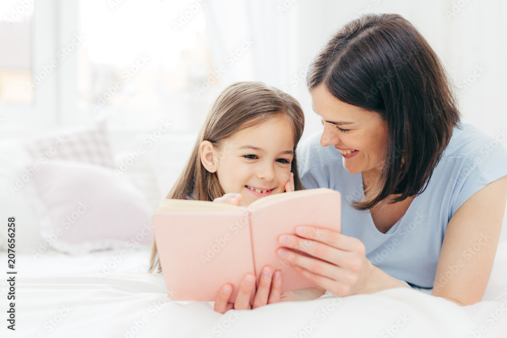 Indoor shot of pleasant looking female child with curious expression, reads interesting book together with her affectionate mother, views colourful pictures, lie on bed in spacious bedroom. Childhood