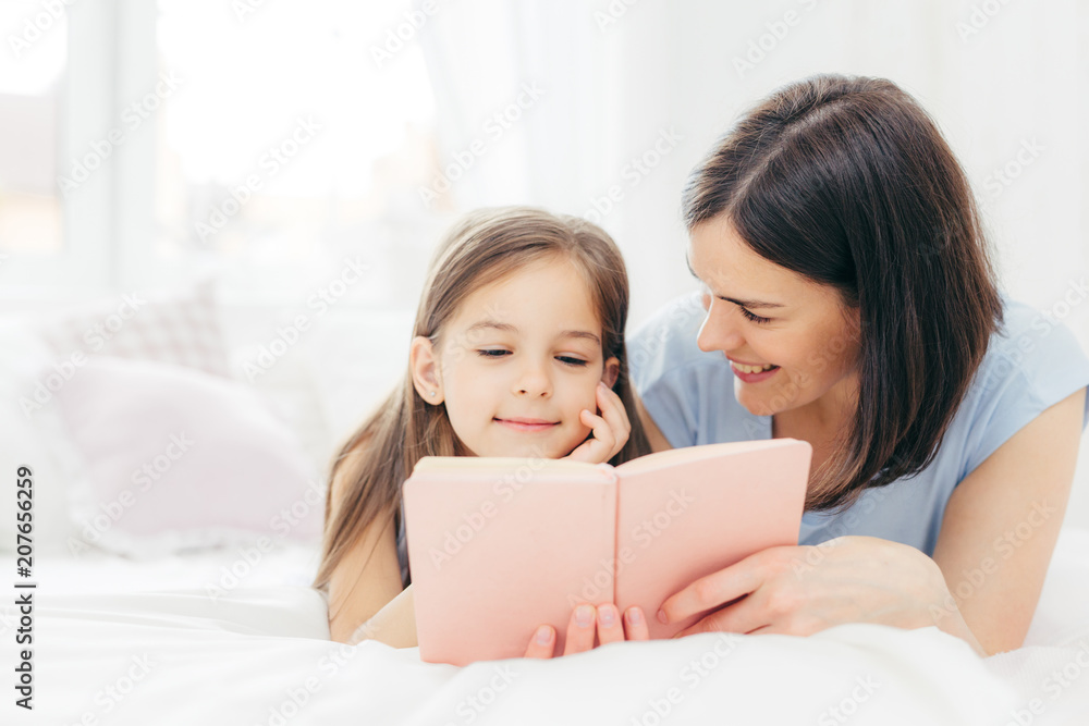 Affectionate young mother with dark hair, looks happily at her small daughter, read interesting book or fairy tale together, enjoy domestic atmopshere at home. People, family, childhood concept Affectionate young mother with dark hair, looks happily at her small daughter, read interesting book or fairy tale together, enjoy domestic atmopshere at home. People, family, childhood concept