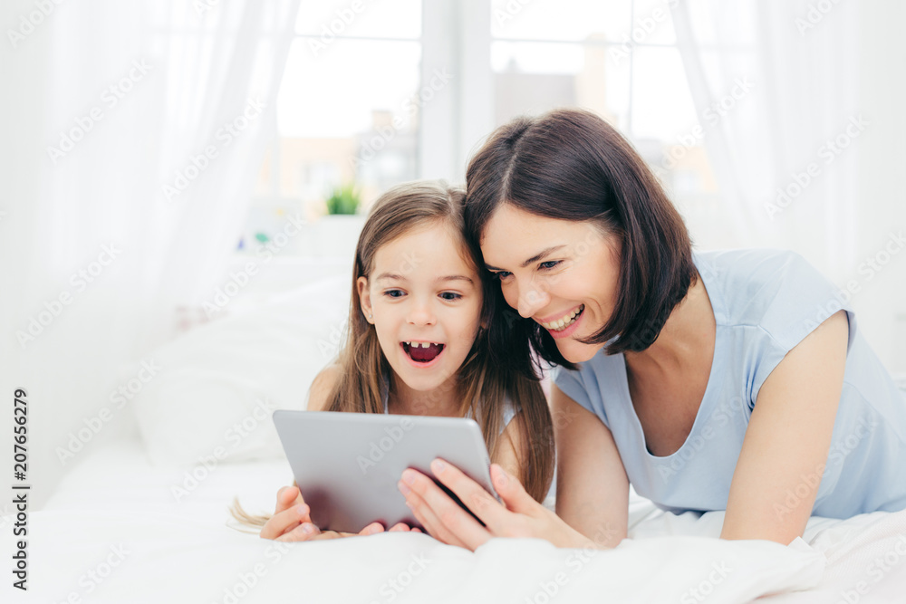 Positive cheerful small female kid and her young mother watch interesting cartoon on tablet, connected to wireless internet, pose against bedroom interior, have happy expressions. Family concept Positive cheerful small female kid and her young mother watch interesting cartoon on tablet, connected to wireless internet, pose against bedroom interior, have happy expressions. Family concept