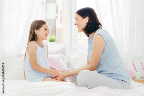 Lovely mother and daughter enjoy togetherness, keep hand together, have pleasant talk, sit on comfortable bed in morning, sit in light bedroom, have good relationships. Family and love concept © VK Studio