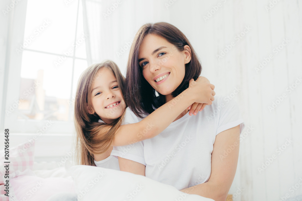 Indoor shot of good looking brunette mother with gentle smile and her small daughter gives hug, enjoy domestic atmosphere, pose against spacious white bedroom interior. Motherhood and family concept Indoor shot of good looking brunette mother with gentle smile and her small daughter gives hug, enjoy domestic atmosphere, pose against spacious white bedroom interior. Motherhood and family concept