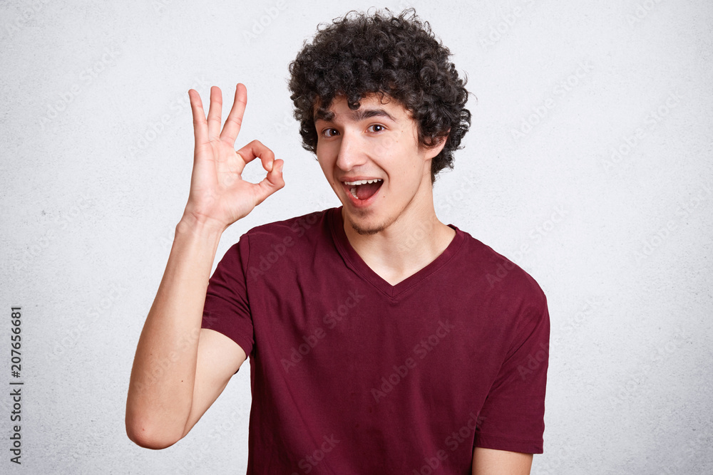 Everything will be fine! Satisfied curly man looks with assured and happy expression, shows ok sign, demonstrates his readiness and awareness, dressed in casual t shirt, isolated over white background Everything will be fine! Satisfied curly man looks with assured and happy expression, shows ok sign, demonstrates his readiness and awareness, dressed in casual t shirt, isolated over white background