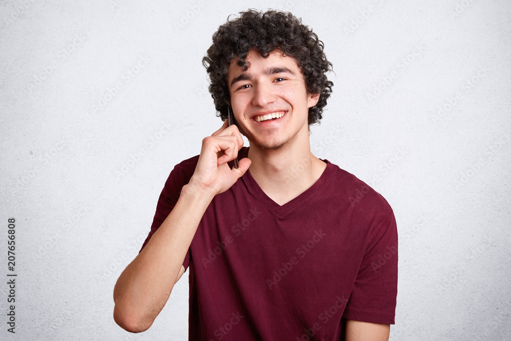 Happy teenager or hipster guy, speaks via cell phone with best friend, has delighted expression, recieves good news, smiles gently at camera, enjoys communication. People and modern technologies Happy teenager or hipster guy, speaks via cell phone with best friend, has delighted expression, recieves good news, smiles gently at camera, enjoys communication. People and modern technologies