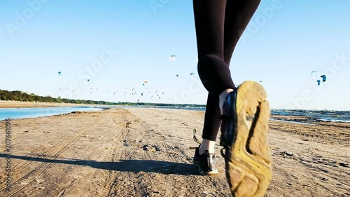 Running female legs in sneakers in the summer close-up against the backdrop of kitesurfers. Woman running in the sea at sunset, slow motion.
