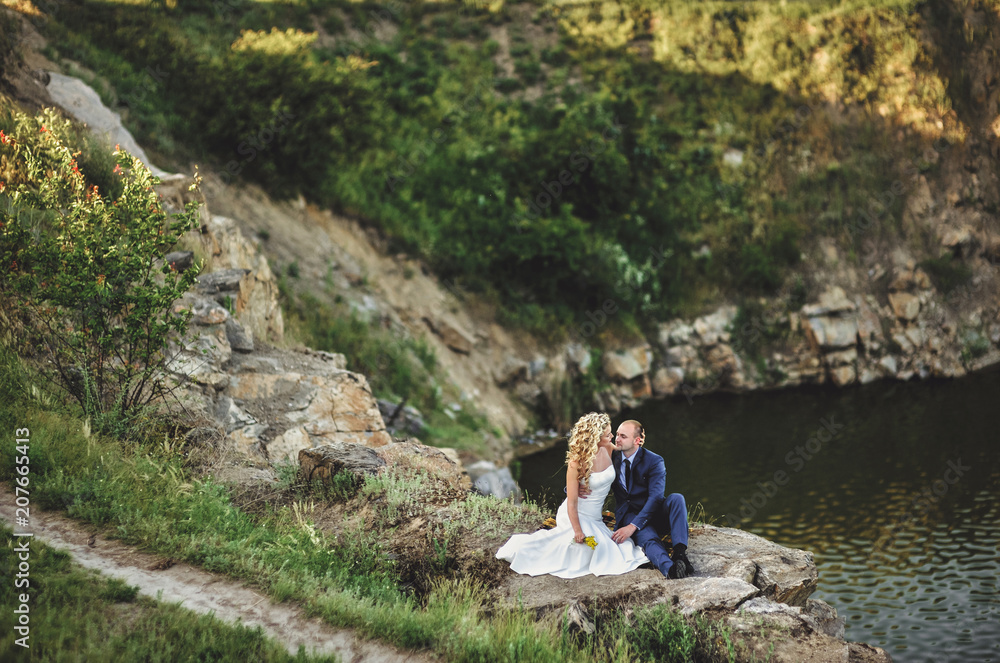 Lovely newlyweds are sitting on a rug and hugging, against the background of the rocks and the river. A stylish bridegroom embraces a beautiful bride with curly hair.