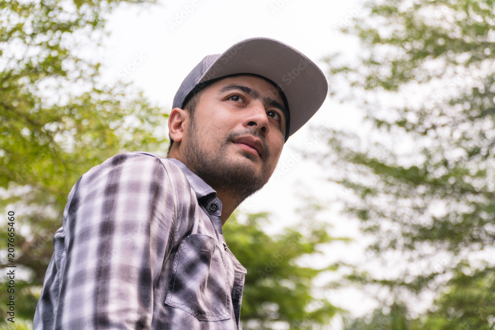 Portrait Handsome man : Good looking young man. He has a beard and wears a hat. He has an interesting smile. Looking for someone. Behind him, full of green trees. Copy space.