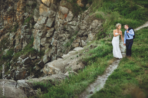 Merry newlyweds go holding hands and laughing, against the background of rocks and green glade. A cheerful groom and a beautiful bride with curly hair walk in the meadow.