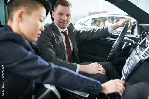 Profile view of pretty Asian potential customer examining new car while sitting on passenger seat, friendly salesman looking at her with warm smile