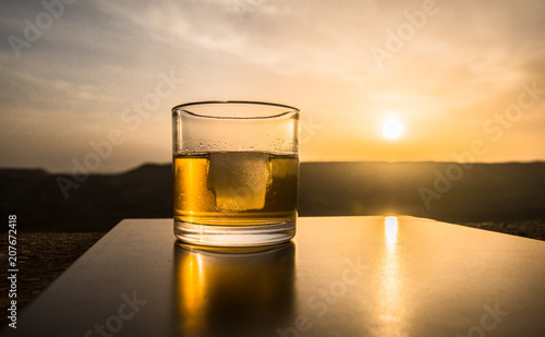 A glass of whiskey with ice on a sunset background or shot of whiskey at sunset dramatic sky on mountain landscape background © zef art