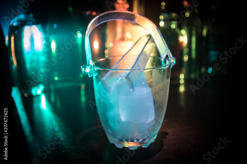 Glass ice bucket on blurred alcohol bottles background with lights and smoke. Club drinks concept. Club bar desk. Ready to serve.