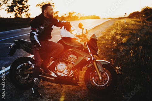 Fototapeta Naklejka Na Ścianę i Meble -  Full length portrait of an awesome young caucasian man sitting on his motorcycle while resting in his vacation trip near the road against sunset.