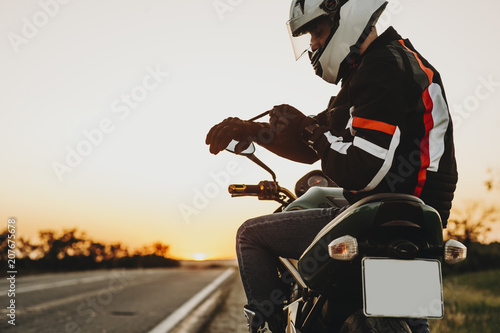 Side view of a caucasian man sitting on the motorcycle and putting his gloves preparing to start his adventure on motorcycle against sunset.