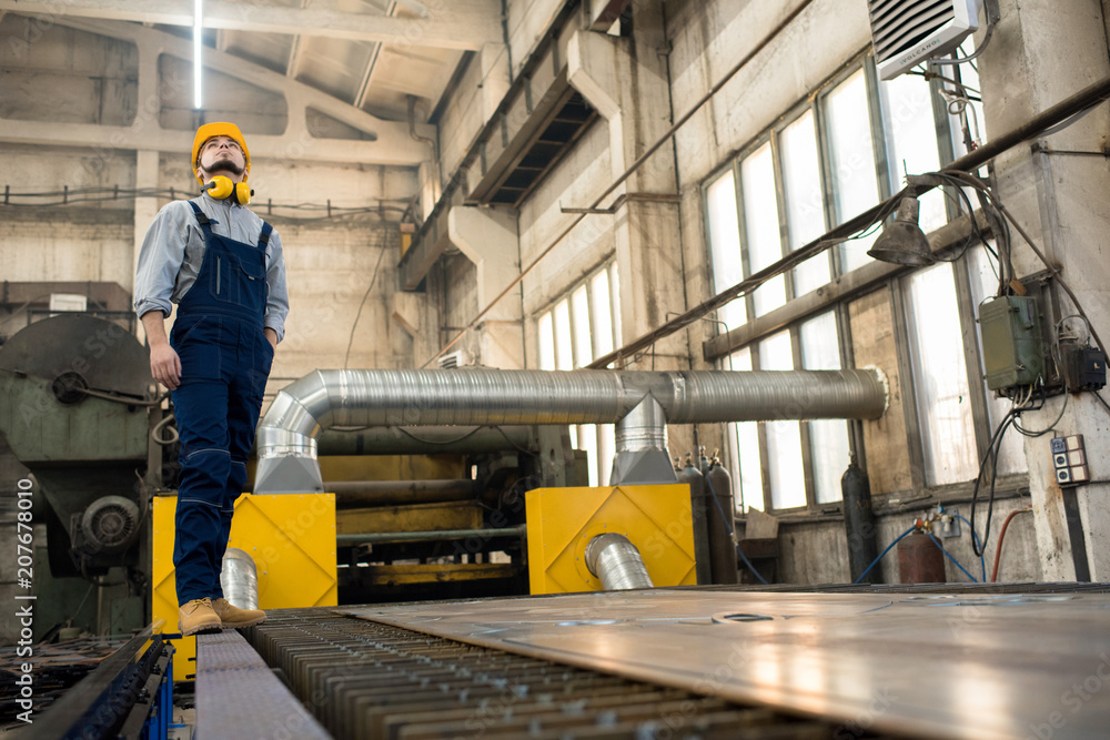 Full length portrait of bearded young technician wearing protective helmet and overall looking upwards with concentration while standing at spacious production department of modern plant