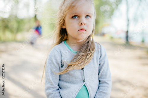 Little adorable beautiful child closeup portrait.  Young small girl with long hair and emotional expressive face mood outdoor summer portrait. Different facial expressions. Childhood.  Baby on road.