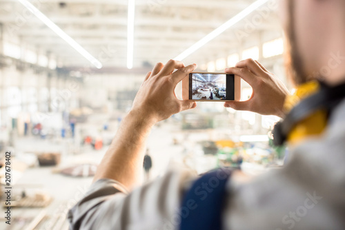 Over shoulder view of unrecognizable inspector wearing ear protectors standing at spacious production department of modern plant and taking picture on smartphone while carrying out inspection.