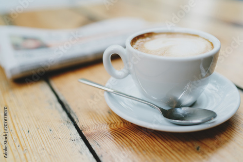 White coffee cup with copy space for the logo, promotional text or design standing on a plate with a spoon. Mockup, drink concept. Hot coffee in a cup standing on a wooden table next to a newspaper.