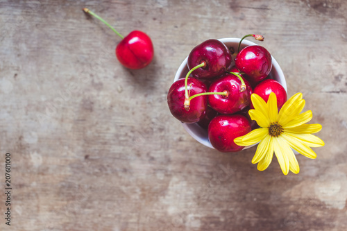 Close up and top view of fresh red cherries decorated with yellow flower in a white cup and a yellow straw on an rustic wooden background. Hello summer, weight loss or healthy diet concept