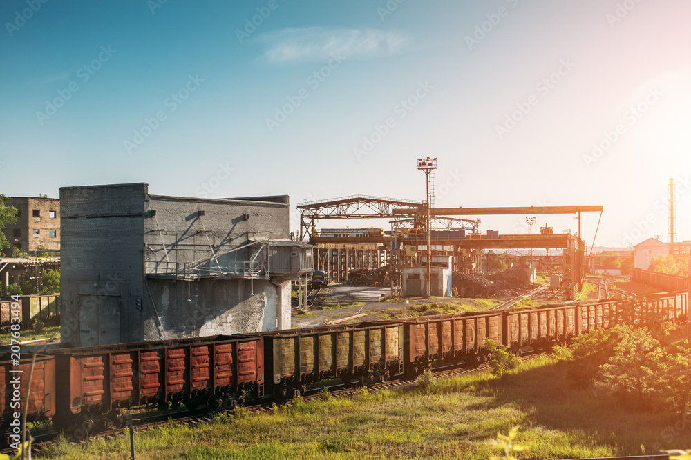 Cargo freight train wagons go on railroad in industrial zone with plants and manufacturing factories of heavy industry at sunset light Cargo freight train wagons go on railroad in industrial zone with plants and manufacturing factories of heavy industry at sunset light