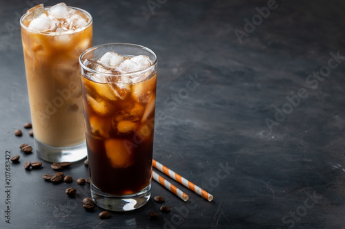 Ice coffee in a tall glass with cream poured over, brown sugar and coffee beans. Cold summer drink on a dark background. With copy space