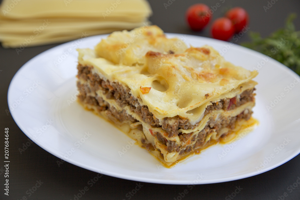 Traditional beef lasagne on a white round plate, black background, side view.