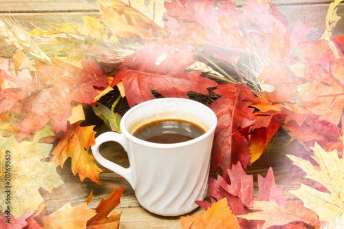 A single hot cup of coffee encircled by leaves in autumn colors on a wooden surface with a texture applied to the fringes of the image.