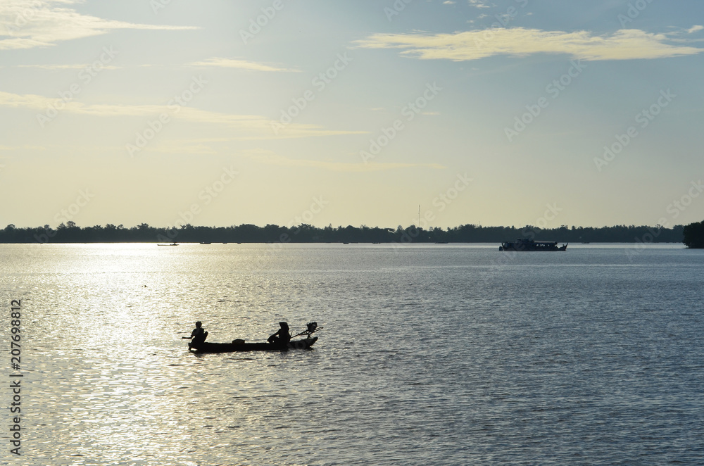 Silhouette of fishermen in a small boat on a river. The afternoon sun has made the water sparkle. Another boat is visible, and a forest covered shore is in the distance.