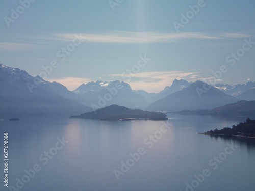 A silver-blue lake surrounded by mountains. Some of the mountains have snow on them. The sun is breaking through the clouds to fall on an island in the lake..