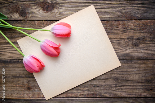 Three pink tulips and a sheet of paper on a wooden background