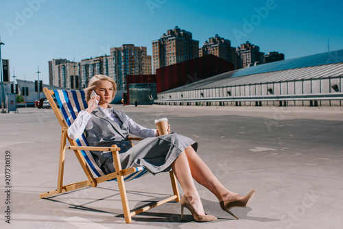 attractive young businesswoman in formal wear sitting in sun lounger with coffee to go and talking by phone on parking