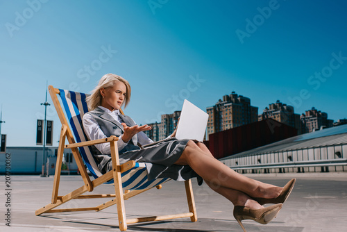 mad young businesswoman in formal wear sitting in sun lounger and looking at laptop on parking