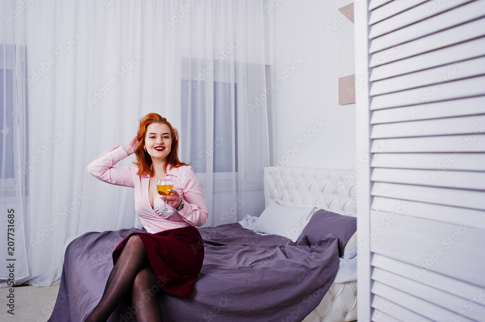 Gorgeous red haired girl in pink blouse and red skirt with glass of wine at hand on the bed at room.