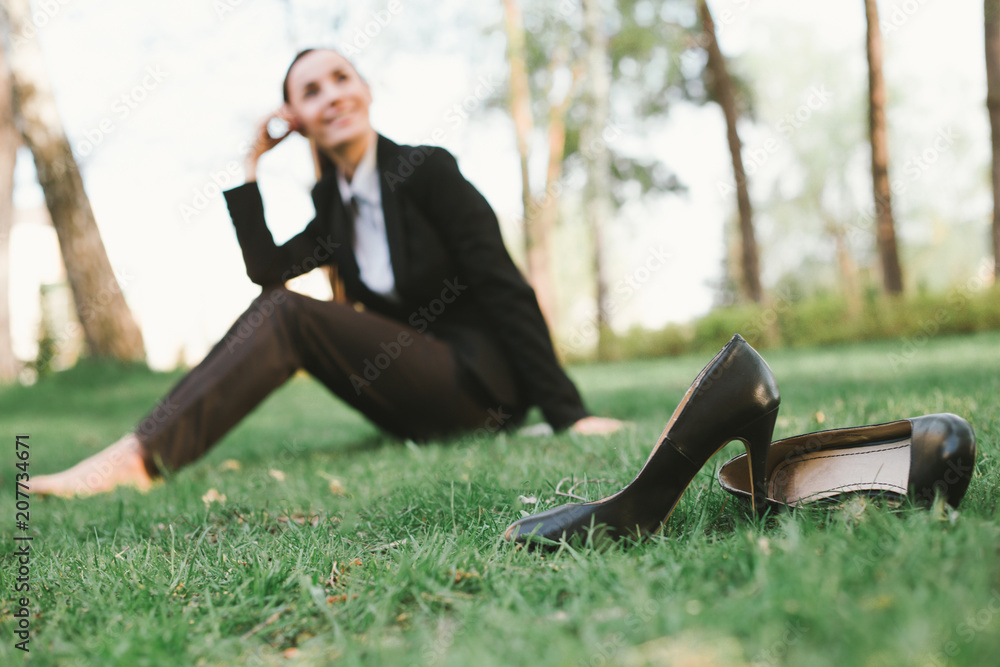 Rest after work in the office. Beautiful young business woman in a black suit is sitting in the park on the lawn and smiling. Women's shoes in the foreground Rest after work in the office. Beautiful young business woman in a black suit is sitting in the park on the lawn and smiling. Women's shoes in the foreground